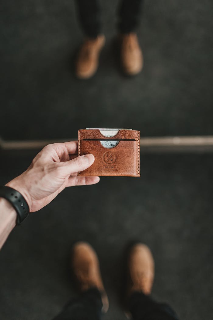 A close-up of a hand holding a brown leather wallet, blurred shoes in the background.
